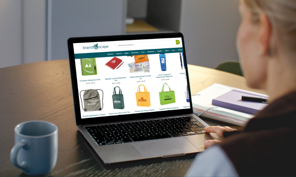 Woman browsing branded merchandise such as bags, notebooks, and tumblers on Brandscape’s website using a laptop.